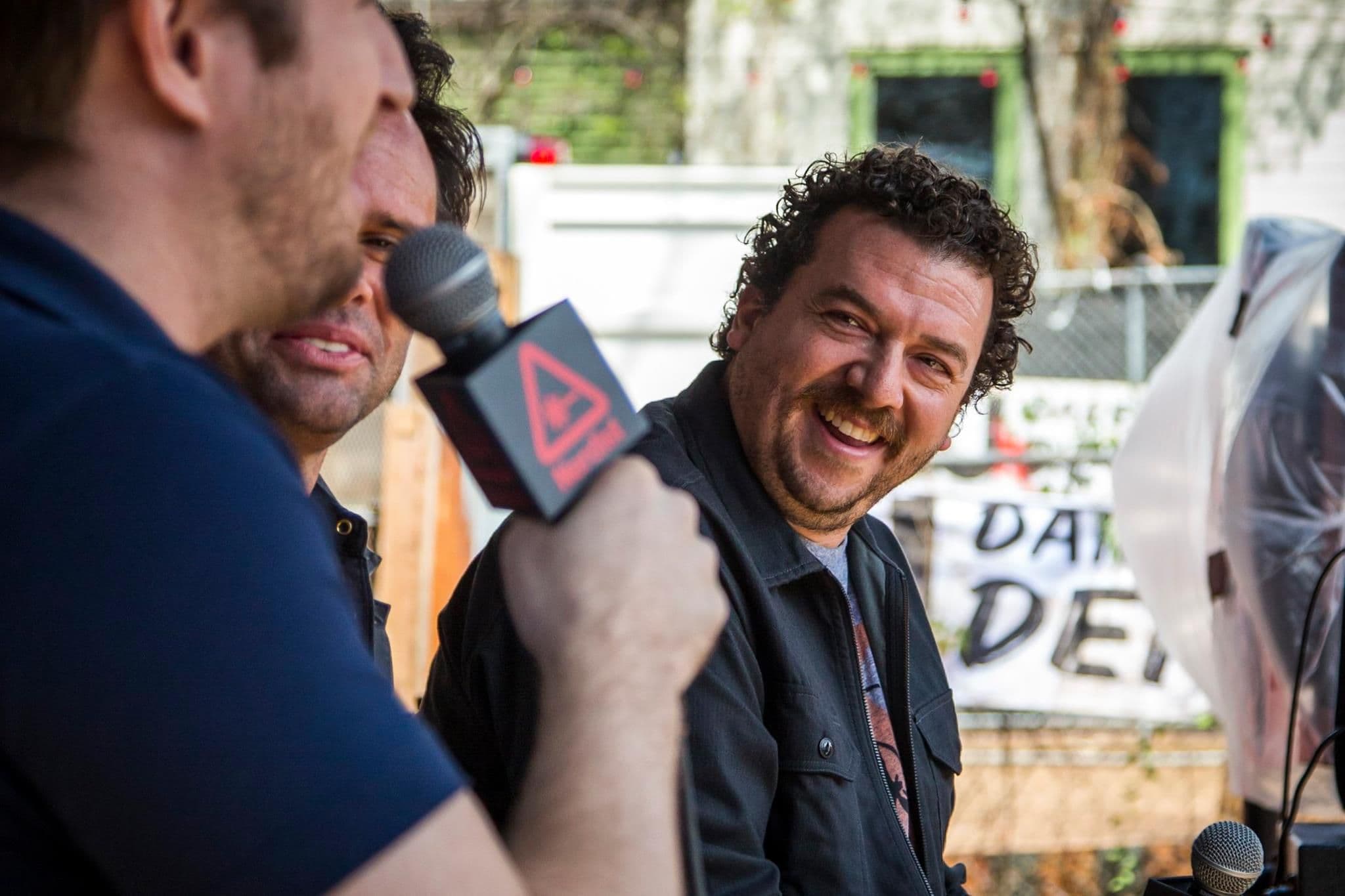 Man laughing during an outdoor interview, with a microphone flag in the foreground and string lights behind a fence
